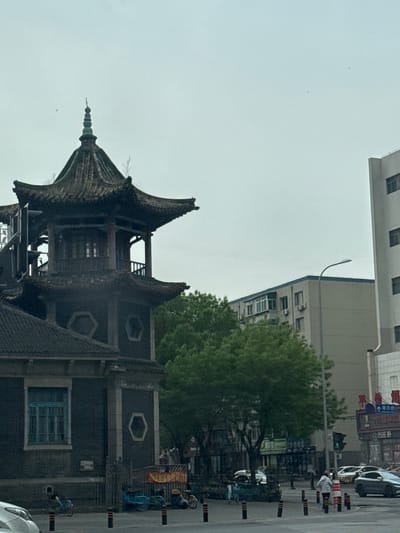 The image shows a traditional Chinese pagoda-style building, likely a temple or historical structure, situated in an urban environment. In the background, there is a modern multi-story building with a street scene visible, including cars, people, trees, street signs, and barriers along the road. Based on the surrounding architecture and streetscape, the image could be from Shenhe District, China, which has a mix of historical sites and modern urban development.