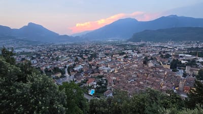 This media file captures an inverted panoramic view of Riva del Garda, Italy, at dusk. The scene is dominated by the town's densely packed buildings with terracotta roofs, cascading down a hillside towards a expansive, tranquil lake. Lush green trees frame the top of the image, suggesting the vantage point is from a higher elevation, possibly a scenic overlook. In the distance, majestic mountains rise, their peaks softly illuminated by the fading light. A thin, elongated cloud, tinged with pink and orange hues, stretches across the sky, indicating the transition from day to night. The sky transitions from a deep blue overhead to a lighter, almost pastel shade near the horizon.

A notable object in the town is a bright blue swimming pool, set within a landscaped area, suggesting a resort or hotel. Streetlights are beginning to glow in the town, hinting at the evening hours. The overall impression is one of serenity and natural beauty, with the man-made town seamlessly integrated into its mountainous and lakeside environment. The time of day is clearly dusk, with the soft, warm light painting the sky and casting long shadows. No people are discernible in the image, and there is no visible text.