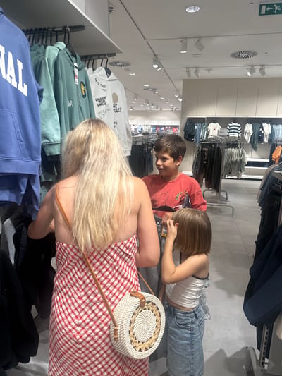 In a clothing store located in the Polígono Industrial De Quart De Poblet, Spain, a woman with long blonde hair, wearing a red and white checkered dress and a round straw purse, stands with her back to the camera. She is looking at clothing items on a rack to her left. To her right, a young boy in an orange t-shirt is looking at something with interest, and a young girl in a white tank top and denim shorts is standing next to him, touching her face. The store environment features clothing racks filled with apparel, track lighting on the ceiling, and a general retail atmosphere.