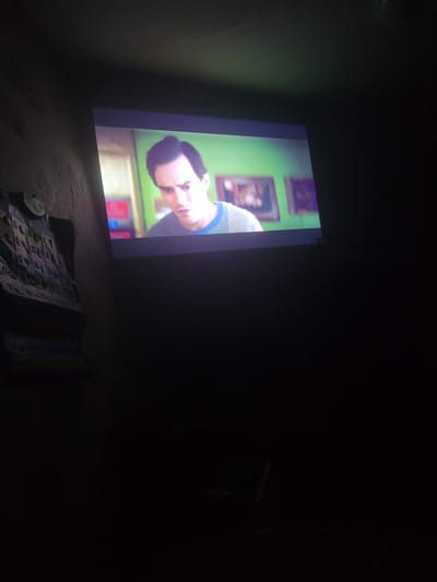 An overhead shot captures a projection screen displaying a close-up of a Caucasian man with dark hair, looking down with a concerned expression. He is wearing a blue and grey striped sweater. The background of the projection shows a green wall with two framed pictures and a blurred interior. To the left of the screen, a wall with a textured pattern is visible, with a metal object and some papers attached to it. The overall environment is dark, suggesting the image was taken in a dimly lit room, likely a home in Hlusk, Belarus, given the context provided. The projector itself is not visible in the frame.
