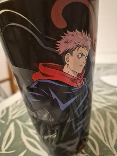 The image shows a close-up of a black cylindrical object, likely a cup or container, with an anime character printed on it. The character has spiky pink hair, sharp eyes, and is wearing a red collar over a dark blue jacket with purple accents. The background is a blurred green and white patterned surface, possibly a tablecloth. The object is positioned on this surface. There is no discernible location in a city.