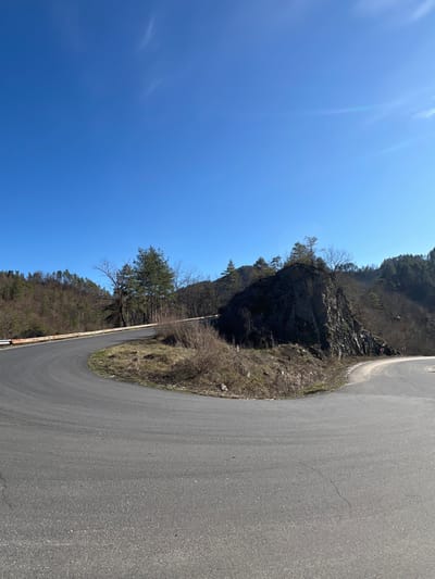 A clear blue sky with faint white contrails dominates the upper portion of the image. The scene captures a winding asphalt road forming a sharp left-hand turn, located near Stoianovo, Bulgaria, in a mountainous or hilly terrain. A secondary asphalt road branches off to the right from the main bend. A weathered, light-colored guardrail, showing signs of rust, lines the outer edge of the left road curve. A prominent dark rock outcrop, topped with evergreen pine trees and sparse bare deciduous trees, is situated to the right of the primary road bend. The surrounding hillsides are covered with a mix of leafless deciduous trees and evergreen conifers, suggesting a late winter or early spring season. The immediate roadside ground cover consists of dry brown grass and sparse shrubs. No individuals or vehicles are present.