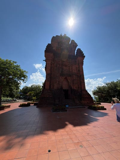 The image shows the Po Nagar Cham Towers, a historical monument located in Tuy Hoa Ward, Vietnam. The central subject is a large, ancient brick tower with a weathered and damaged upper section. The tower is constructed from dark red bricks and features a prominent arched entrance with steps leading up to it. Sunlight streams down from the upper right corner of the image, creating a lens flare and casting long shadows on the tiled ground in front of the tower.

The ground is paved with large, reddish-orange square tiles. Several rectangular flower beds with red flowers are strategically placed around the base of the tower and in the foreground. Lush green trees frame the scene on the left and right sides, with a glimpse of distant hills or mountains in the background. The sky is a vibrant blue with scattered white clouds, indicating a clear, sunny day.

In the foreground on the right side, a person, viewed from behind, is taking a photograph of the tower with a smartphone. The person is wearing a white shirt and what appears to be blue shorts. There are no other visible people or significant interactions taking place. The overall environment suggests a well-maintained historical site visited by tourists.
