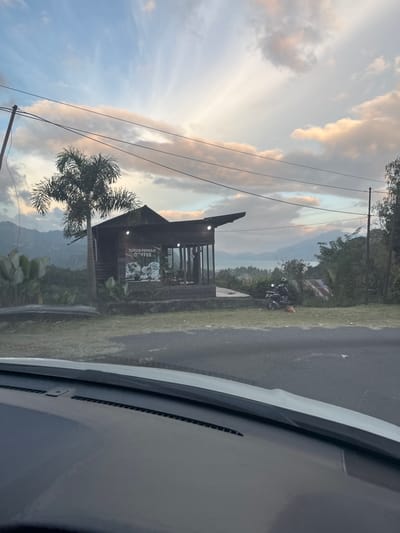 The image depicts a scenic view from a vehicle's dashboard, looking out at a small wooden cafe situated on a hillside. The cafe, labeled "TOTOR PEMULO COFFEE," has large windows and an overhang. A palm tree stands to the left of the cafe, with lush green foliage and distant hills in the background. The sky is a mix of blue and clouds, with hints of sunset colors. A motorcycle is parked near the cafe, and a cat is resting on the grass. The foreground shows the interior of a car, with its dashboard visible. The location is specified as Payatumpi Baru, Indonesia.