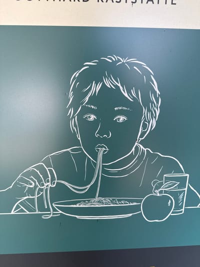 This is a photograph of a sign in Schattdorf, Switzerland, featuring a white line drawing of a child eating spaghetti. The child is depicted with their mouth open, slurping a strand of pasta from a fork. In front of the child is a bowl of spaghetti, an apple, and a glass of liquid. The background is a solid teal color. The words "GOTTHARD RASTSTÄTTE" are visible at the top of the sign, indicating it is located at a rest stop on the Gotthard Pass.
