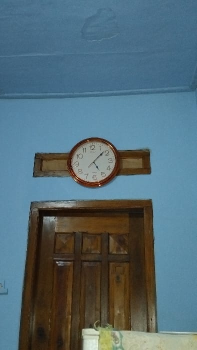 The image shows a clock mounted on the wall of a room in Panjalu, Indonesia. The clock is round with a copper frame and has black hands pointing at 11:50. The clock is mounted on a brown wooden shelf. To the right of the clock is a wooden door. The walls of the room are light blue. There is a white ceiling with a light blue stain. The door is open to reveal a room, which is not visible. The coordinates provided (latitude: -7.142, longitude: 108.278) indicate that this location is in the city of Panjalu, Indonesia.