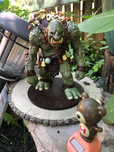 An orc-like figurine, painted green with various warts and growths, stands on a circular wooden base. The figurine is adorned with mushrooms and small skulls on its back and shoulders, and a brown loincloth. It has large, yellowed teeth and is wearing dark brown armbands and leg wraps. In front of the orc, on a larger, rough-hewn log base, is a small, solar-powered toy bear with a Canadian flag decal on its chest. To the right of the orc, a small green frog figurine is perched on the edge of the log. The scene appears to be outdoors, with lush green foliage and a wooden fence in the background. The lighting suggests it is daytime. The location is identified as the city of Severn, Canada.