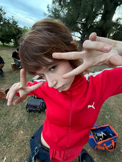 A person in a red Puma jacket makes a V-sign with both hands, obscuring their face slightly. They have brown hair and freckles. The setting appears to be outdoors, with a grassy area, trees, and a partly cloudy sky visible. There's a blue and orange bag, a camera case, and what looks like a wooden crate or bench nearby. The image has been rotated counter-clockwise.