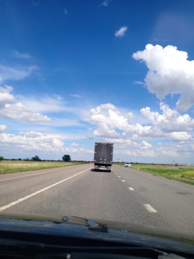 The image shows a highway scene on a bright, cloudy day, possibly taken from the perspective of a driver. A large truck with a dark trailer is in the right lane, traveling away from the viewer. Further down the road, a white car is visible. The highway is bordered by dry, grassy fields with scattered trees. The sky is a vibrant blue, dotted with fluffy white clouds. The scene appears to be in a rural area, potentially in or near Voznesenovka, Russia, given the vegetation and the style of the road and vehicles. The asphalt road has visible white lane markings. The foreground shows the dashboard and windshield of a vehicle, with a wiper and a reflection of the sky visible.