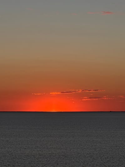 The image depicts a sunset view in Marina di Ravenna, Italy. A fiery orange and red glow dominates the sky near the horizon, transitioning to softer orange and grey hues higher up. The sea, appearing as a textured grey surface, occupies a significant portion of the right side of the image, with a stark contrast against the vibrant sky. The sun is either setting just below the horizon or obscured by a thin layer of clouds, creating a diffused, radiant light. There are faint, vertical cloud formations highlighted in orange near the sunset. The scene is devoid of people or other explicit subjects; the focus is on the natural elements of the sunset and sea.