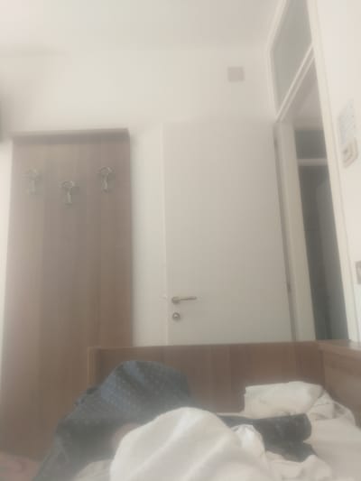 The image depicts an indoor scene, likely a hotel room in Lignano Sabbiadoro, Italy. The room appears to be furnished with a wooden wardrobe or closet featuring three metal hooks, a door with a silver handle and lock, and a bed covered with white and dark patterned bedding. In the foreground, there is a white blanket or sheet. The overall lighting is soft, suggesting ambient indoor light or possibly an overcast day. The room is tidy, with no visible clutter or disarray. The presence of a wardrobe, bed, and door suggests a typical lodging environment.