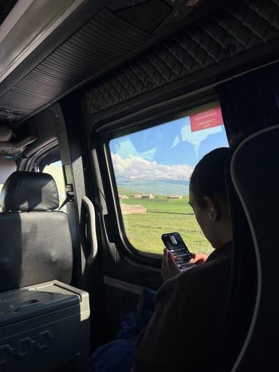 This media file captures an interior view of a vehicle, likely a tour bus or van, looking upwards and towards the front. The dominant subject is a young person, seen from behind and slightly to the side, engrossed in their smartphone. They are wearing dark clothing and appear to have an earbud in their left ear.
The setting is the interior of the vehicle, with elements like quilted black upholstery on the ceiling, a sunroof, and black leather seats visible. A gray handle is attached to the ceiling. Outside the sunroof, a bright, sunny sky with fluffy white clouds is visible, suggesting daytime. The scene outside the vehicle is a picturesque rural landscape with green fields and some distant structures that appear to be ancient ruins. The sun is high in the sky, casting strong shadows within the vehicle.
A notable detail is a rectangular red sign affixed to the sunroof, with text in both Georgian and English. The English text clearly reads "EMERGENCY EXIT". The person's focus on their phone suggests a moment of leisure or entertainment during travel, possibly during a scenic drive. The overall mood is relaxed and focused on personal engagement amidst a journey through a visually appealing landscape. The location is identified as Gegharot, Armenia.