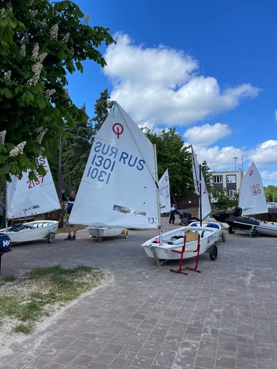 The image captures an outdoor scene at what appears to be a sailing club or boat yard in Pionersky, Russia, under a bright blue sky with scattered white clouds. Several small white sailboats, likely Optimist dinghies, are lined up on a paved surface.
In the foreground and mid-ground, there are multiple boats on individual trolleys or dollies with wheels, some red, some metallic. The largest and most prominent boat in the foreground has a white sail displaying the red "Q" logo, "20Я RUS", and the numbers "1301" (and its mirror image "10ει"). Another boat to its left shows "S" and "2137" on its sail. Further back, another boat's sail has the "Q" logo and numbers "80" and "080", while another sail shows "Q" and "180". A small outboard motor is visible on the stern of one boat. A blue bag with "HH" (Helly Hansen) is next to the foreground boat.
Two individuals are visible among the boats, appearing to attend to them. One person is standing near a boat in the middle-right. The ground is paved with rectangular grey and brown bricks, with a narrow strip of sand and sparse green grass along the right edge. In the background, green trees, including some with white blossoms (possibly chestnut trees), and a multi-story building with white-framed windows are visible. The scene suggests preparations for sailing or boats being stored on land during daytime.