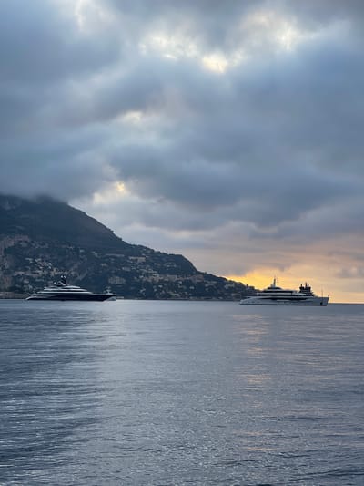 The image depicts a coastal scene near the city of Èze, France, under a cloudy sky at what appears to be dawn or dusk. Two large yachts are present on the water. The yacht on the left is dark-hulled with a white superstructure and is closer to the mountainous coastline, which is dotted with buildings. The yacht on the right is predominantly white with multiple decks and is further out in the water, positioned against a background of a softly illuminated sky where the sun is peeking through the clouds. The water is calm, with gentle ripples reflecting the subdued light. The overall atmosphere is tranquil and majestic, highlighting the opulence of the yachts against the natural beauty of the Mediterranean coast.