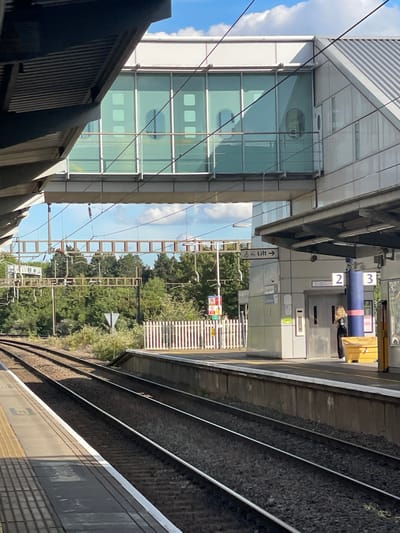 This is an eye-level, medium shot of a train station in Luton, United Kingdom. The scene is oriented vertically, with the tracks running diagonally from the foreground to the background, suggesting a slight downward angle. Sunlight streams down, indicating it is daytime with clear skies and fair weather.
The foreground features a train platform with dark gray, textured surfaces and yellow markings. Next to it, parallel train tracks are visible, filled with dark gravel and gleaming steel rails. To the left, a modern building with a glass facade and horizontal louvers dominates the left side of the frame. The building's reflective windows offer glimpses of interior structures.
In the middle ground, a walkway or bridge extends across the tracks, connecting to the building. A woman with blonde hair is partially visible near a lift entrance, indicated by a sign with a directional arrow and the word "Lift." Above the lift, numbers "2" and "3" are displayed, possibly indicating platform or level numbers. Other signs with text are present but difficult to decipher.
The background is filled with lush green trees, suggesting a natural element within the urban environment. Overhead, electrical wires and support structures for the train line are visible against a bright blue sky with scattered white clouds. The overall impression is one of a functional, modern transportation hub set against a backdrop of nature on a sunny day.