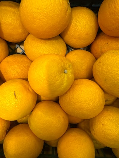 This is a close-up, top-down photograph of a large pile of bright yellow oranges, filling the frame. The oranges are round and have a textured, dimpled rind. They are densely packed together, creating a visually appealing mound of citrus fruit. Some oranges are partially obscured by others, emphasizing the abundance. The lighting appears bright, highlighting the vibrant color of the fruit. There is no visible text, people, or specific indications of time of day or weather. The setting appears to be a market or grocery store, suggested by the way the oranges are piled and the dark background which could be a crate or shelf. The image conveys a sense of freshness and natural bounty.