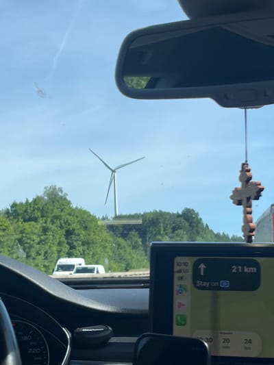 The image captures a view from inside a car looking out through the windshield. In the foreground, the dashboard is visible, with a portion of the speedometer showing speeds up to 240. A car navigation system displaying a map and route information is mounted on the dashboard. A hanging wooden cross is suspended from the rearview mirror. In the mid-ground, a wind turbine stands tall against a clear blue sky with some wispy clouds. Lush green trees form a dense backdrop behind the wind turbine and on a gentle slope. Two white vans are parked or stopped on a road in the distance, partially obscured by foliage. The scene appears to be set on a sunny day. The location is identified as Icking, Germany.