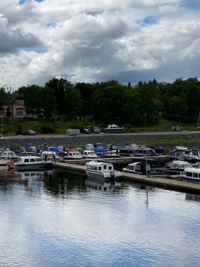 The media file shows a row of boats docked along a pier on a body of water. The boats are varied in size and color, with many appearing to be modern motorboats. Some have enclosed cabins, while others are open. Several boats are white, and some have blue or orange accents. A few people are visible on the decks of the boats, suggesting they are either arriving, departing, or simply enjoying a day out.
The setting is a marina, with a tree-lined shore visible behind the pier. The trees are lush and green, indicating it is likely summer or late spring. The water is calm and reflects the sky, which is partly cloudy with patches of blue visible. The overall atmosphere appears peaceful and relaxed. A small building, possibly a house or part of the marina facilities, is visible near the shore.
The time of day is difficult to ascertain precisely due to the cloud cover, but the lighting suggests it is daytime, perhaps late morning or afternoon. There are no overt signs of specific events, but the presence of multiple boats and people suggests a leisurely activity, such as boating or fishing. No significant text is visible in the image. The location context provided is Sortavala, Russia, which aligns with a coastal or lakeside setting.