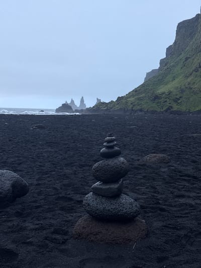 A black sand beach scene in Vik, Iceland, dominated by a stack of rocks arranged linearly from small to large, leading away from the viewer. The beach stretches towards distant rock formations in the ocean. To the left, a moss-covered cliff face looms. The sky is overcast. There are no people visible.