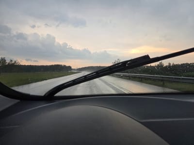 The image, taken from inside a vehicle, shows a wet highway in Gudulinė, Lithuania, under a cloudy sky with hints of sunset. The windshield is partially obscured by a wiper blade, and there are visible raindrops on the glass. The road ahead is slick and reflects the overcast sky. On either side of the road, there are lines of trees, and a metal guardrail is visible on the right. The foreground features the dark interior of the car, likely the dashboard. The overall scene suggests driving in rainy or damp conditions during twilight.