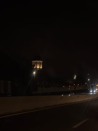 A nighttime scene in Jilava, Romania, captures a distinct, brightly illuminated octagonal building with a dome-like roof, identified as the Jilava Prison water tower, elevated against a dark sky. The structure is bathed in warm yellow light. In the immediate foreground, a dark asphalt road surface features a solid white lane marking on its right edge. A light-colored concrete barrier runs parallel to the road on the left. Multiple artificial light sources are present across the midground, including a glaring white streetlight positioned slightly below the illuminated building, other scattered amber and white lights, and headlights from several vehicles on a road further to the right. The overall environment is dark, with no visible human activity or interactions.