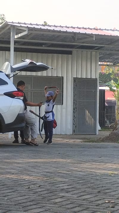In Trosobo, Indonesia, a young boy, wearing a blue cap, white shirt with blue trim, and dark pants, stands on a paved area, holding a dark red strap above his head with both hands. He appears to be participating in some form of activity, possibly martial arts training, as he is also wearing a dark belt with a sheath containing a curved blade. Beside him, a man in a black shirt and light-colored pants, wearing flip-flops, leans against the open trunk of a white SUV. The man holds a dark object, possibly a phone, in his right hand. The scene unfolds in front of a building constructed with corrugated metal siding and a metal roof. A doorway with decorative metalwork is visible on the right side of the building. To the left, the white SUV is parked. The background features foliage, suggesting an outdoor setting.