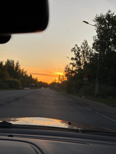 A photograph taken from inside a vehicle shows a road at sunset in Pontonny, Russia. The sky is filled with vibrant orange and yellow hues, indicating the sun is low on the horizon. Trees line both sides of the road, silhouetted against the colorful sky. Streetlights are visible along the road, with one illuminated. A yellow triangular road sign depicting a pedestrian crossing is present on the right side of the road. The asphalt road shows signs of wear and tear, with some potholes and faded white lane markings. The hood of the car, with a windshield wiper, is visible in the foreground, reflecting the sunset.
