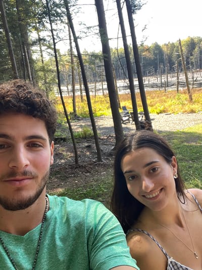 This image captures a close-up selfie of two young adults outdoors in Otego, United States. On the left, a light-skinned male with dark curly hair and a short beard wears a green short-sleeved shirt and a dark beaded necklace. On the right, a light-skinned female with long dark straight hair smiles, wearing a light-colored, possibly checkered, spaghetti-strap top, a silver necklace with a small pendant, and a silver earring. Both subjects are looking directly at the camera.
The background reveals a natural environment. Immediately behind the individuals are several tall, slender trees with dark trunks, some coniferous. The ground beneath the trees consists of green grass and patches of lighter-colored soil or gravel. In the mid-ground, a body of water or marshland is visible, characterized by numerous standing dead tree trunks. Beyond this marsh, a dense line of live trees with mixed green and autumnal foliage is present under a bright, clear sky. Two dark outdoor chairs and a small pile of logs are visible in the mid-ground among the trees and ground cover.