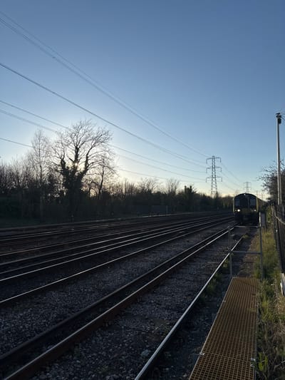 The image depicts a multi-track railway corridor within the city of Southampton, UK. This configuration of numerous parallel tracks suggests an area near a major railway junction, a sidings facility, or an approach to a traincare depot, such as those found around Northam or Millbrook.
The scene is viewed at a low sun angle, indicating either early morning or late afternoon/early evening, with the sun partially obscured by bare deciduous trees on the left, silhouetting them against a clear blue sky. Approximately eight to nine parallel railway tracks, composed of steel rails on ballast, recede into the distance from the foreground.
An electric multiple unit (EMU) train, identifiable by its yellow-fronted livery and illuminated headlights, is visible on the rightmost set of tracks, approaching the viewer from the distant horizon. Overhead power lines, including smaller traction lines directly above the tracks and larger transmission lines supported by substantial lattice-style electricity pylons further to the right, extend across the sky, running parallel to the railway. A utility pole stands on the far right.
In the immediate foreground on the right, a section of a yellowish-brown metal grate platform or walkway is visible, positioned alongside a metal fence that separates the viewing point from the tracks. The left side of the tracks is bordered by a dense line of bare deciduous trees and undergrowth. No people are visible in the scene.