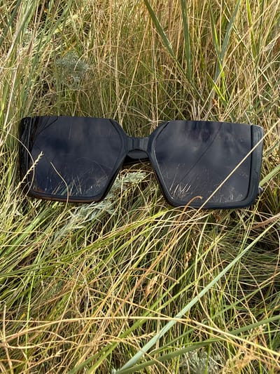 A pair of black, oversized sunglasses is lying on the ground amidst tall, dry grass. The sunglasses have a thick, angular frame and dark, reflective lenses. The lenses show scratches and reflections of the surrounding vegetation. The grass is a mix of green and golden-brown hues, appearing dry and wind-swept. A small, pale grey plant is visible among the grass near the left lens of the sunglasses. The scene suggests an outdoor environment, possibly a field or roadside in a rural area. The provided metadata indicates the location is in the city of Өндіріс, Kazakhstan.