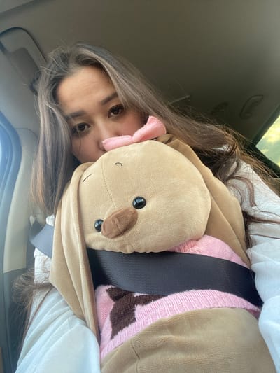 A young woman is inside a vehicle, holding a stuffed animal. The stuffed animal appears to be a brown bunny with a pink sweater and a pink bow on its head. The woman has long brown hair and is looking towards the camera. A seatbelt is visible across the stuffed animal and the woman's chest. The background suggests the interior of a car with a window visible on the right side. The image is a close-up selfie.