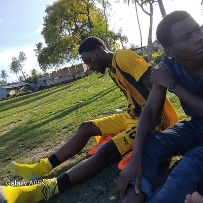 Two young men are sitting on a grassy field in Nungwi, Tanzania, located at latitude -5.726 and longitude 39.301. The man on the left is wearing a yellow and black soccer jersey, orange and black soccer cleats, and yellow socks. The man on the right is wearing blue jeans, a black and white shirt, and a black watch on his right wrist. He is holding the left arm of the man on the left. The man on the left is looking down at his left leg, and the man on the right is looking at the camera. There are trees and buildings in the background. The scene appears to be taken during the day in a sunny location. The grassy field appears to be a soccer field. A "Galaxy A05s" watermark is visible in the lower left corner of the image.