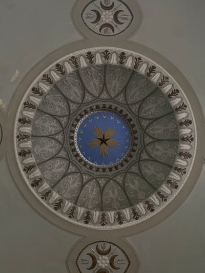 The image captures an ornate ceiling inside a building in Schwetzingen, Germany. The ceiling is primarily a pale green color and features a large circular design at its center. The design includes concentric rings of decorative patterns, including stylized leaves and floral motifs in shades of white and brown. At the very center of the circle is a blue disk with a gold star and smaller white floral elements. To either side of the central circle are smaller circular medallions, each containing crescent moon shapes, arrows, and a central white flower. The overall impression is of a historically significant, well-preserved interior space.