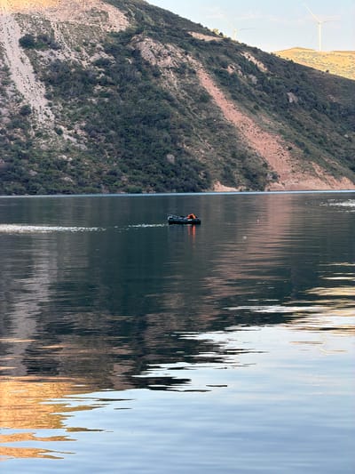 A serene lake reflects a mountainous terrain covered in sparse vegetation and soil. A small inflatable boat with an orange-clad individual is positioned in the center of the lake, creating ripples on the water's surface. In the background, two wind turbines are visible on a distant hilltop. The image appears to capture a tranquil moment in a natural landscape, with the boat and its occupant adding a focal point to the scene. The location, Brichmulla, Uzbekistan, is known for its picturesque natural beauty, which is evident in this photograph.