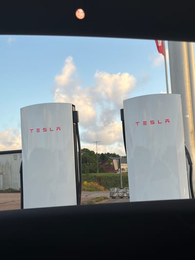The image shows a Tesla Supercharger station in Hogstorp, Sweden, under a cloudy sky. Two white Tesla charging stations are visible in the foreground, each with the red Tesla logo. In the background, there is a building with a red roof, some trees, and a flagpole with a red and white flag. The ground is paved with asphalt and there are some yellow flowers. The image appears to be taken from inside a vehicle, looking out through the windshield. A blurry baseball or similar object is visible in the upper center of the image, possibly a reflection or something on the windshield.