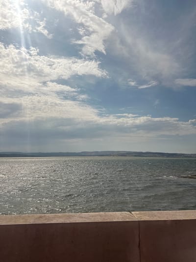 The image shows a panoramic view of the sea and sky, with a concrete barrier in the foreground. The sea is choppy and reflects the sunlight, creating a shimmering effect. The sky is partly cloudy, with rays of sunlight breaking through the clouds. In the distance, a hazy shoreline with rolling hills can be seen. The scene appears to be located along the coast of Narbonne, France, likely near the Canal de la Robine or the Mediterranean Sea. There are no people or objects of significant note, other than the natural elements and the man-made barrier. The overall impression is one of a serene and natural landscape.
