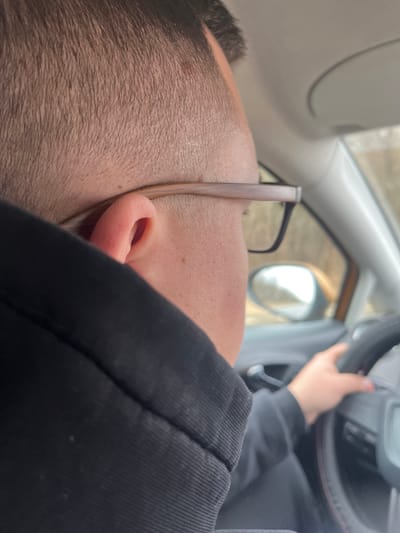 A man with dark hair and glasses is lying down inside a car. His head is near the driver's side window. A hand is visible on the steering wheel. The car's side mirror is visible in the background through the window. The interior of the car is light gray. The man is wearing a black hooded jacket. The location of Rogachovo, Russia cannot be determined from the provided image.