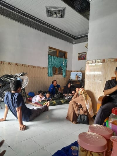 The image shows a living room in a home located at -7.637 latitude and 112.141 longitude in the Kecamatan Purwoasri area of Indonesia. The room features a white ceiling with black and grey trim, white walls, tiled floors, and a large window with a turquoise curtain. A motorcycle is partially visible in the left foreground, near a man seated on the floor. He is wearing a dark blue shirt and dark pants, and has his legs extended in front of him. To his right, a young girl in a pink shirt lies on a blue blanket on the floor, while another child, a boy in a blue shirt, is seated on a white cushion, looking at a small screen. Further in the background, a man wearing a green shirt and brown pants sits on a cushion, looking at the two children. He has grey hair. To his right, a woman with long dark hair is sitting on a cushion. She wears a brown and white long-sleeve dress.  Next to the woman is a figure obscured by a brown and white cloth, possibly another individual.  Behind the group sitting on cushions is a television screen, showing a woman in a long dress.  A large object resembling a chair is located in the right foreground.  Behind the chair is a man seated, wearing a blue long-sleeve shirt and dark pants. He is looking at a large jar with a blue lid, positioned on the floor in front of him. The jar appears to be filled with an unknown substance.  In the right foreground, there are two jars with pink lids, which appear to contain some sort of snack.   The scene suggests a family gathering or a group of people enjoying snacks and leisure time.