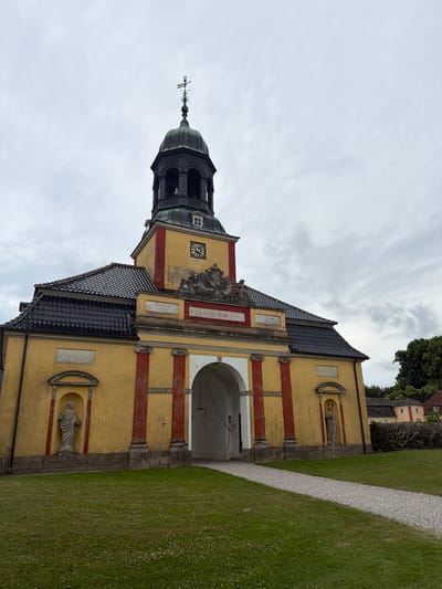 The image shows the exterior of a yellow building with red trim, possibly a church or historical building, located in Allerslev, Denmark. The building features a dark, domed tower with a weather vane and a clock face. Niches with statues are visible on the facade, and an arched entryway leads into the building. The building is surrounded by a well-maintained grassy area with a gravel path. In the background, trees and another building with a red roof are visible. The sky is overcast, suggesting a cloudy day. No people are visible in the image. The overall impression is one of historical architecture and serene surroundings. The time of day is indeterminate due to the overcast sky. There is no visible text in the image, apart from numerals on the clock face, which are not clearly legible.