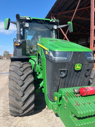 A green and yellow John Deere 8R series tractor is parked outdoors in Krushovo, Bulgaria, under a clear blue sky. The tractor is oriented with its front-right section facing the viewer. The front features a black grille with a prominent John Deere logo, flanked by large rectangular headlights and additional work lights mounted on the cabin roof and front fenders. A large, deeply-treaded front tire is visible on the left side. An exhaust pipe extends upwards from the left side of the engine compartment. The interior of the cab is partially visible through tinted windows, showing a steering wheel and controls. A heavy, multi-ribbed green front weight block is attached to the tractor's front hitch. A red cylindrical fire extinguisher is mounted horizontally on top of this front weight block. The ground beneath the tractor is dry, unpaved, light brown dirt or gravel. In the immediate background to the right, a large structure with exposed reddish-brown steel support beams and girders is visible, consistent with a large agricultural shed or industrial building. Distant trees and a smaller, older building are visible in the far left background. Strong sunlight illuminates the scene, casting defined shadows.