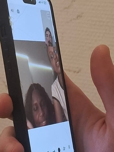 A person holds a black smartphone with a cracked screen. The screen displays a video call with two participants. One participant is a light-skinned individual with glasses, visible in a smaller window. The second participant, in a larger window, is a dark-skinned person with curly hair wearing a white shirt. The background is a white wall with some shadows. The person holding the phone has fair skin and a visible fingertip.