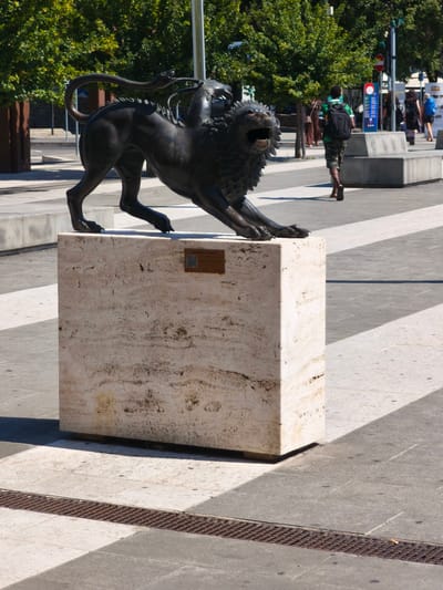 The image shows a bronze sculpture of a chimera, a mythical creature, with a lion's body, a serpent's tail, and a goat's head emerging from its back. The sculpture is positioned in a public square in Arezzo, Italy, indicated by a plaque on its pedestal that reads "CHIMERA di AREZZO" and features a QR code. The chimera is depicted in a dynamic pose, with its mouth open and forelegs extended, appearing to be in motion. It rests on a large, light-colored stone pedestal.
The square has a paved surface composed of large, light-colored square tiles intersected by darker gray tiles and a metal grate running horizontally across the foreground. The scene is bathed in bright sunlight, casting strong shadows from the sculpture and other elements in the environment.
In the background, several people are visible. A young man with a backpack is walking away from the camera, and two women are walking in the distance. Trees with green foliage line the edge of the square, and there are also concrete benches and planters. Traffic lights and signs are present on a pole in the background, suggesting a street or intersection nearby. The overall impression is of a sunny day in an urban public space.