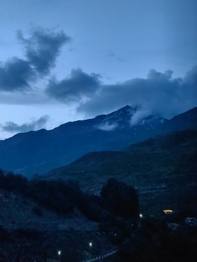 The image depicts a serene mountain landscape in the city of Gushal, India, likely during twilight or pre-dawn hours. The sky is a deep blue, with dramatic, dark clouds creating a sense of impending weather or the fading of daylight. A prominent mountain range dominates the middle ground, with its peaks partially obscured by swirling mist and clouds. Snow-capped portions are visible on the highest summit, hinting at the elevation and cooler temperatures. The slopes of the mountains are rugged and appear to be covered in vegetation, though the low light conditions make details indistinct. In the foreground, the terrain is darker and more densely vegetated, with trees and shrubs casting deep shadows. A few artificial lights, likely streetlights or lights from buildings, are visible in the distance, suggesting human habitation within the otherwise wild environment. These lights are small and scattered, emphasizing the vastness of the natural landscape. The overall impression is one of natural beauty and a tranquil, remote setting.
