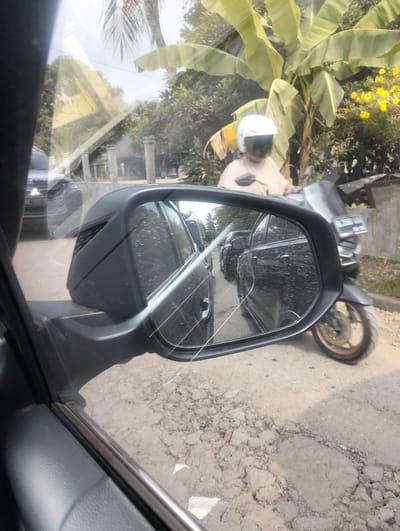 The image captures a scene from the perspective of a car driver in Teupinreudeup, Indonesia, looking into the side mirror and out the window. In the side mirror, two black cars are visible in traffic, with raindrops on the mirror's surface. Outside the car, a person wearing a white helmet and a light-colored shirt rides a motorcycle. Behind the motorcycle rider, a banana tree with large green leaves and yellow flowers is prominent. The ground is a dirt road with patches of asphalt. The sky is overcast. The overall environment suggests a rural or semi-urban setting.