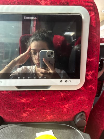 The image shows a reflection of a person taking a selfie in what appears to be a screen or a window of a public transport vehicle. The person, with dark, curly hair and wearing round-framed glasses, is holding a light-colored smartphone (likely an iPhone given the dual camera setup) to capture the image. A design or sticker is visible on the back of the phone case. The person is wearing a light-colored top with some textured detailing and a bracelet on their left wrist.
The immediate setting around the reflective screen is characterized by vibrant red fabric upholstery with a black speckled or patterned design, typical of public transport seating. The screen itself has a white/light gray bezel. To the right of the screen, a black plastic fixture, possibly part of the seat or armrest, is visible, along with a small yellow object partially peeking into the frame.
The activity is the person taking a mirror selfie. The lighting suggests it is daytime. A notable detail is the mirrored text "SIMROLL" (appearing as "110RWI S" due to reflection and image orientation) visible on the screen's surface in the reflection. The overall scene suggests the person is traveling in a bus or train in Bayrampaşa, Turkey.