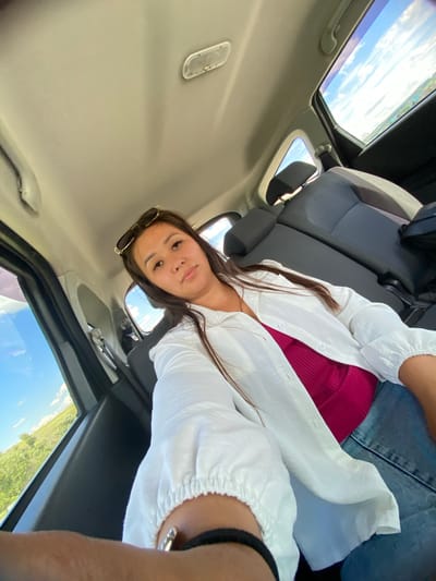 A woman is sitting in the back seat of a car, taking a selfie. She has long, dark hair and is wearing a white open shirt over a pink top and jeans. Sunglasses are perched on her head. The car interior is visible, including the seats, ceiling, and windows. Outside the windows, a bright blue sky with white clouds and green landscape can be seen, suggesting it is daytime and possibly a rural or suburban setting. The image was captured in Yuldybayevo, Russia.