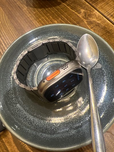 The image shows a silver Apple Watch with a black and silver band in a speckled blue ceramic bowl. A silver spoon rests across the top of the bowl. The watch display shows a number pad, indicating a passcode is required. The bowl sits on a wooden surface with visible wood grain. The location is not discernible from the image. The only city/region in the UK mentioned is Mole Valley, which is likely where the image was taken, but the environment is not specified.