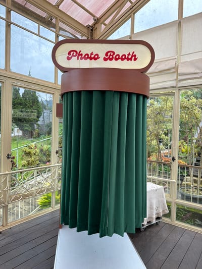 The image shows a photo booth inside a glass structure, possibly a greenhouse or conservatory. The sign on the photo booth reads "Photo Booth" in red lettering. The booth is surrounded by dark green curtains. Through the glass walls, one can see outdoor foliage, suggesting a garden setting. The location appears to be Candikuning, Indonesia, based on the reflection of "Kebun Raya Eka Karya Bali" which is the name of the Botanical Garden, that is located in Candikuning.