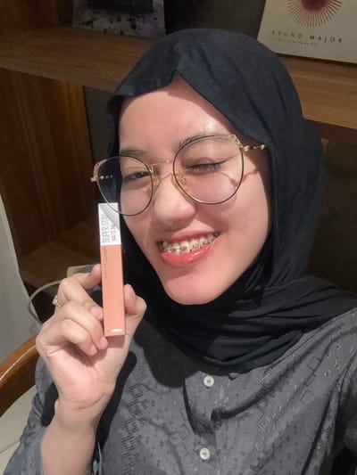 A young adult female is depicted in a close-up, chest-up shot, indoors in Patrang, Indonesia. She wears a black hijab, gold-framed eyeglasses with decorative temples, and a grey patterned long-sleeved shirt with white buttons. Her teeth display visible dental braces. She is winking with her right eye (viewer's left) and smiling. Her right hand, wearing a ring on the ring finger, holds a light peachy-orange rectangular cosmetic product package. Visible text on the package includes "MAYBELLINE NEW YORK" and "SUPER STAY MATTE INK". The background features wooden shelving with a partially visible framed item containing the text "BRUNO MAJOR" and a red circular design.