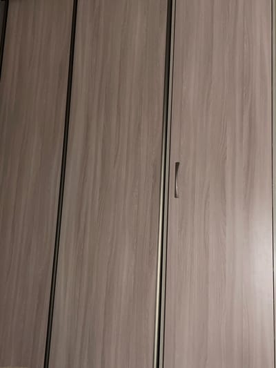 This is a close-up, head-on shot of a wooden dresser with three drawers. The wood has a light grey-brown, weathered look with a visible grain pattern. Each drawer is separated by a thin, dark horizontal line, likely a seam or integrated handle design. The top drawer has a single, curved, dark metal handle positioned centrally. The lighting suggests an indoor setting, possibly with artificial light, as there are no strong shadows or indications of natural light sources. The overall impression is one of a functional piece of furniture in a neutral, possibly domestic, environment. No people are visible, and no text is present. The scene is static, with no activity taking place.